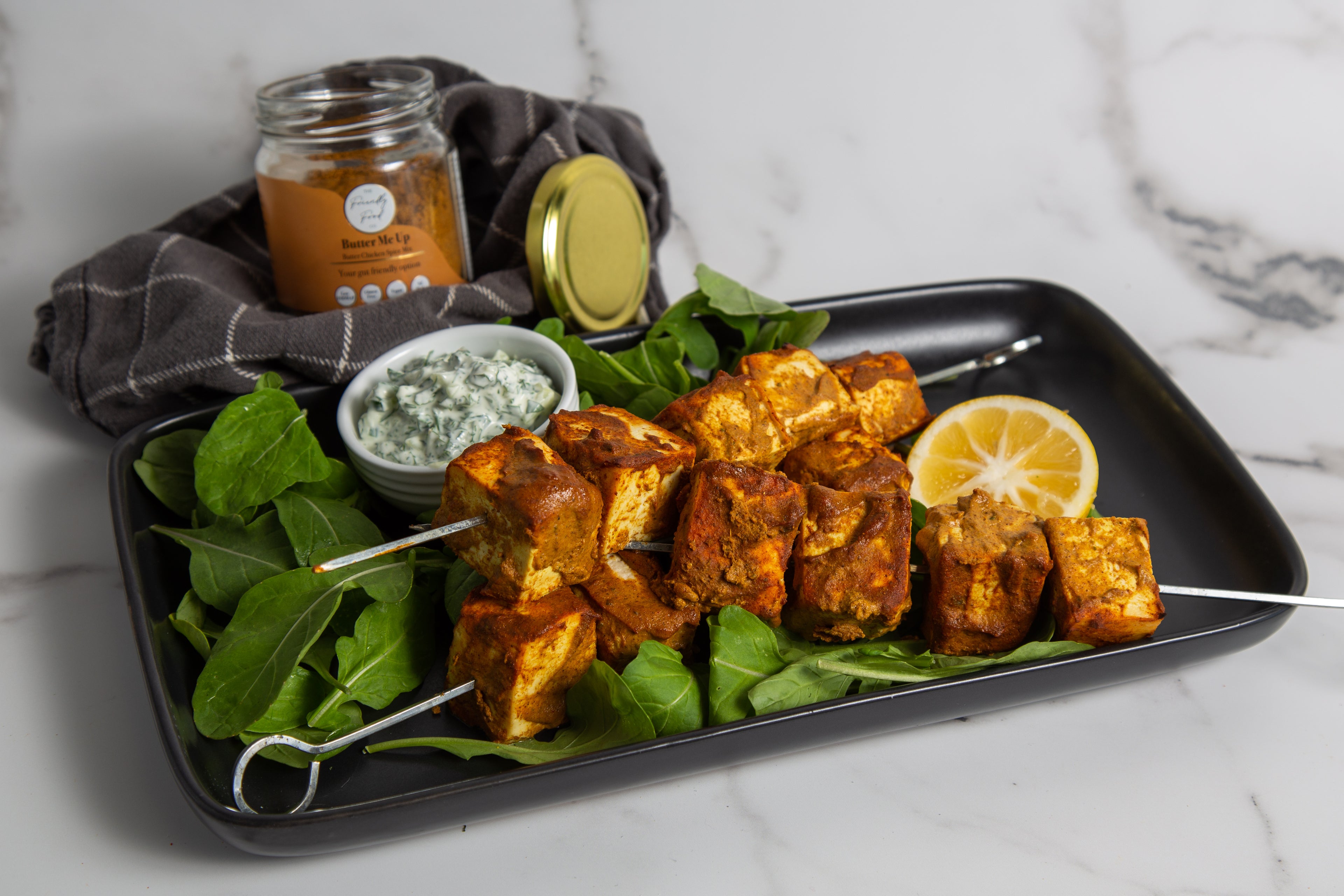 Platter of Butter Ne Up Marinated Paneer Skewers served with lemon, rocket, and herby cucumber yoghurt sauce on a marble surface.