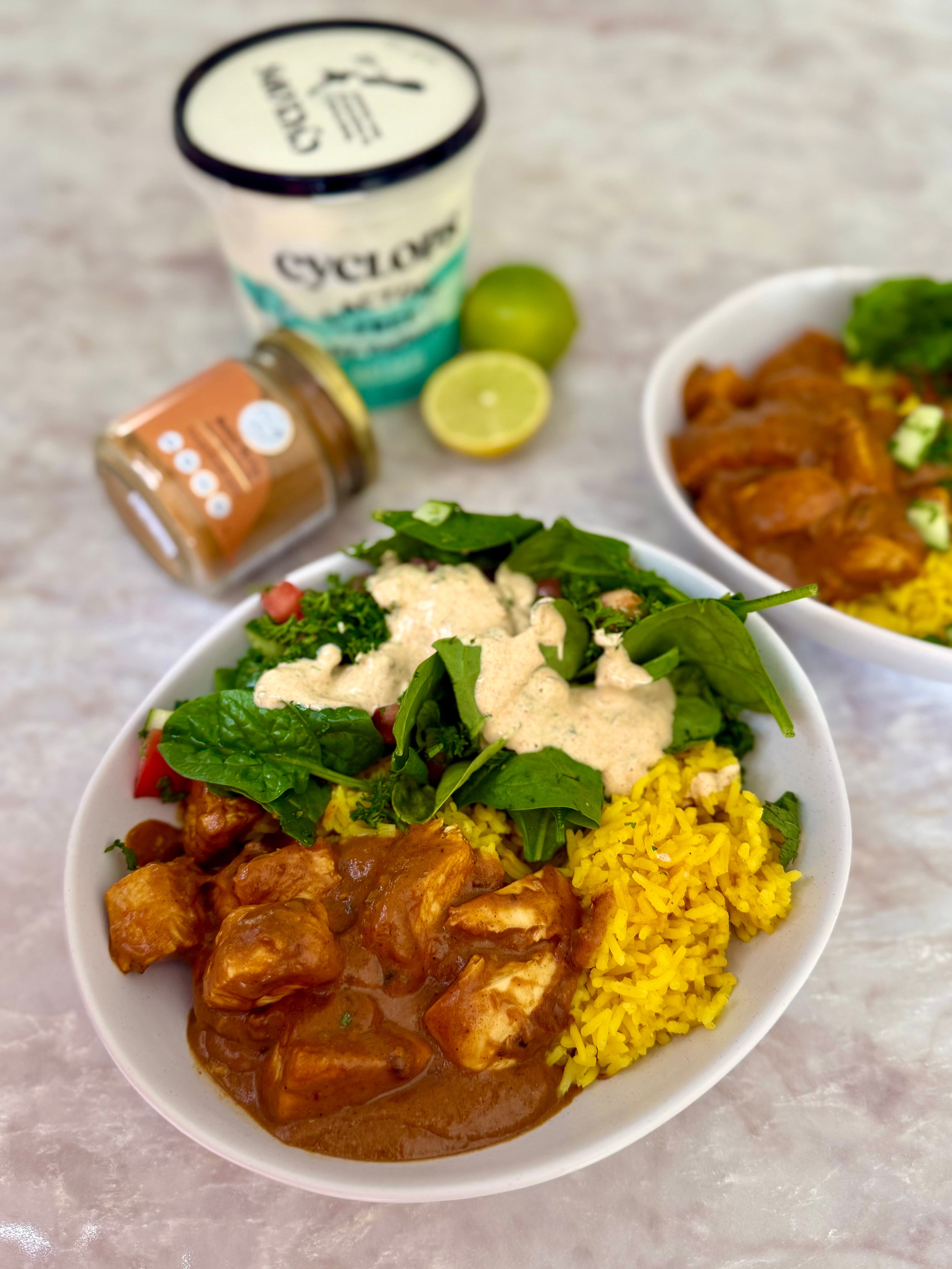 Plate of butter chicken curry with cumin, turmeric rice and salad, with a container of yogurt and jar of The Friendly Food Co Butter chicken spice in the background.