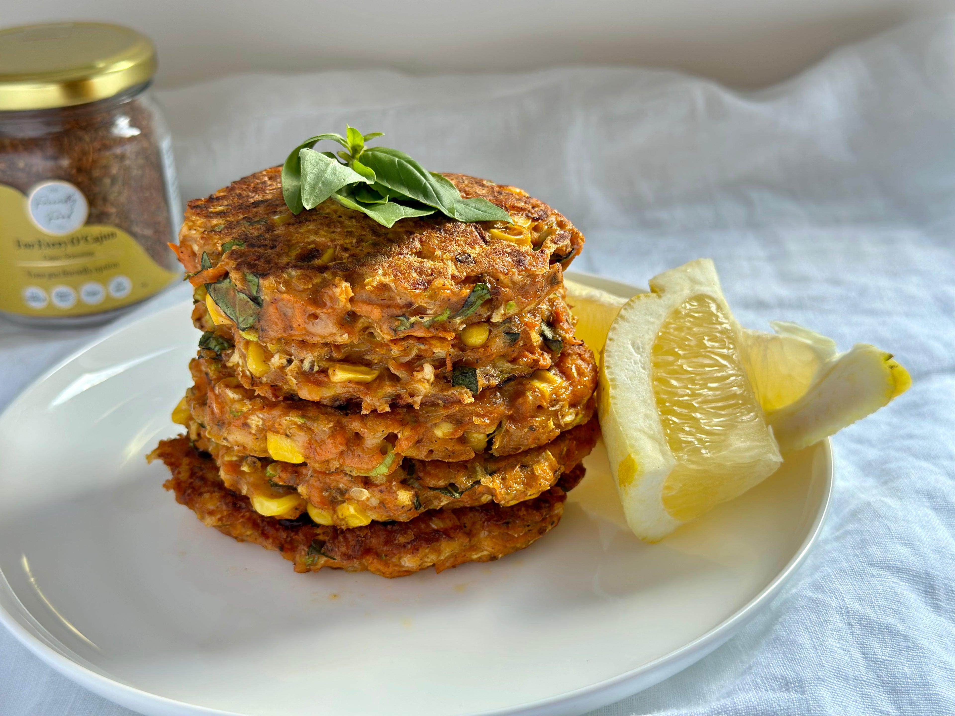 Stack of Cajun, Courgette, Carrot & Corn Fritters on a plate with lemon wedges and a jar of The Friendly Food Co Cajun seasoning in the background.