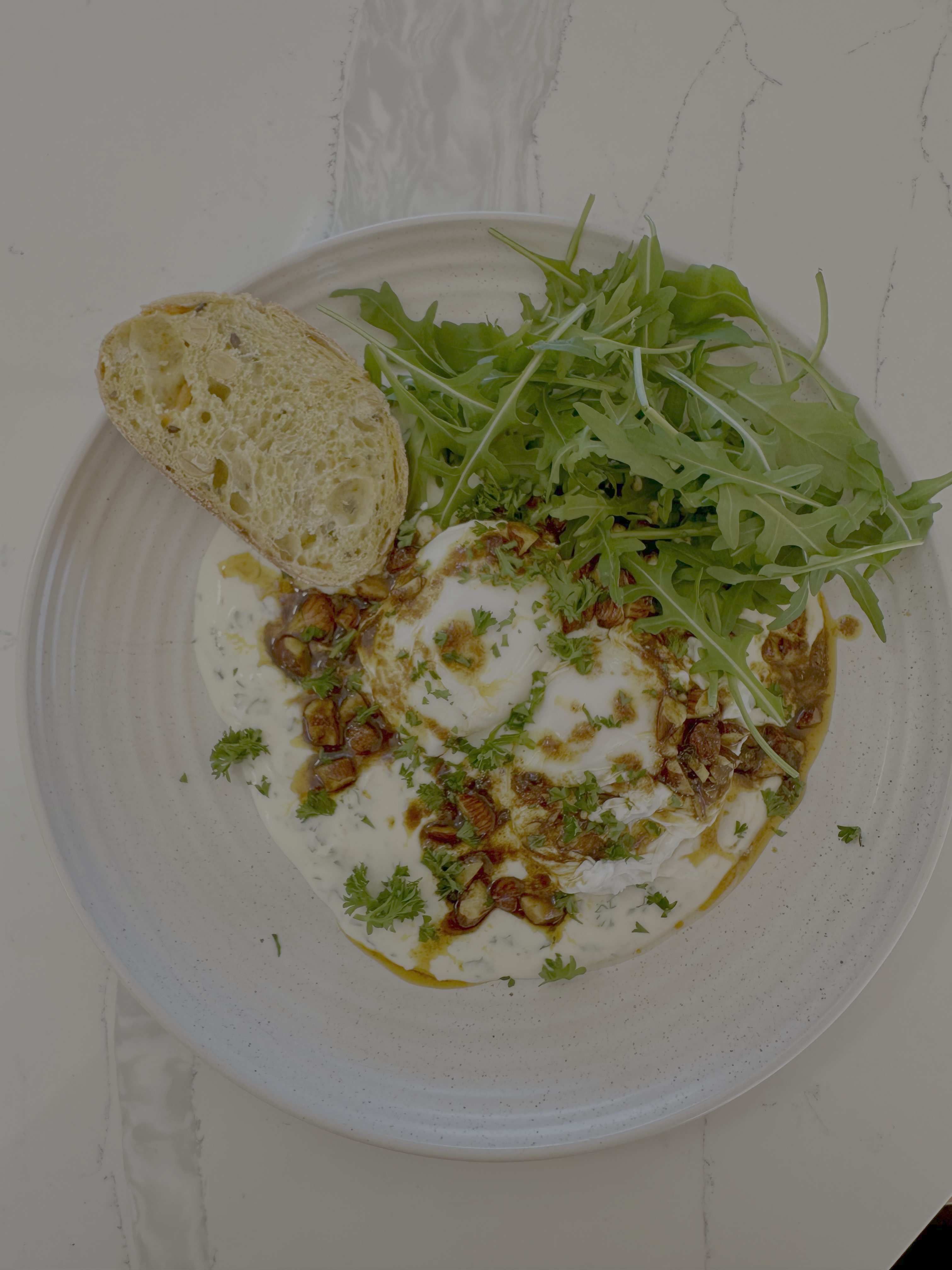 A plated food dish featuring poached eggs in a creamy white sauce topped with crispy brown bits and herbs, served with a slice of toasted bread and fresh rocket on a speckled white plate over a light marble surface.