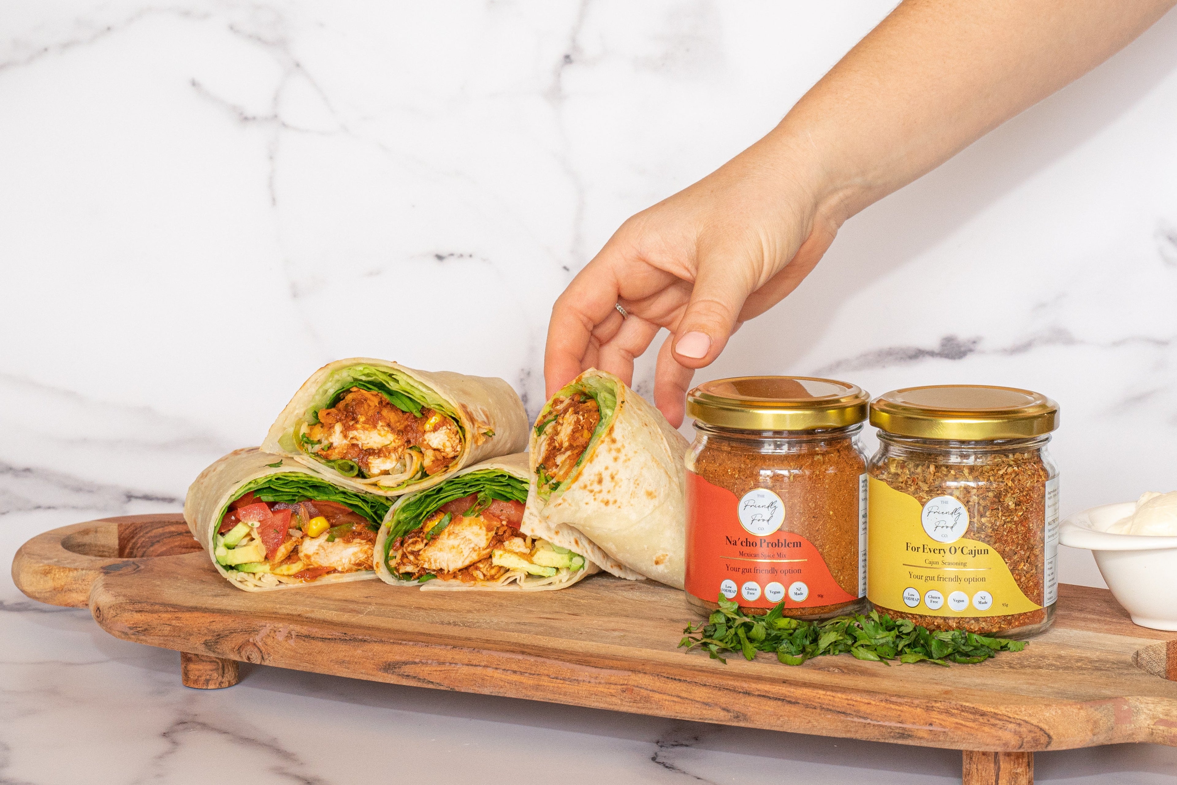 Hand reaching for a Low FODMAP Chicken Burrito on a wooden board with The Friendly Food Co. spice mixes on a marble background