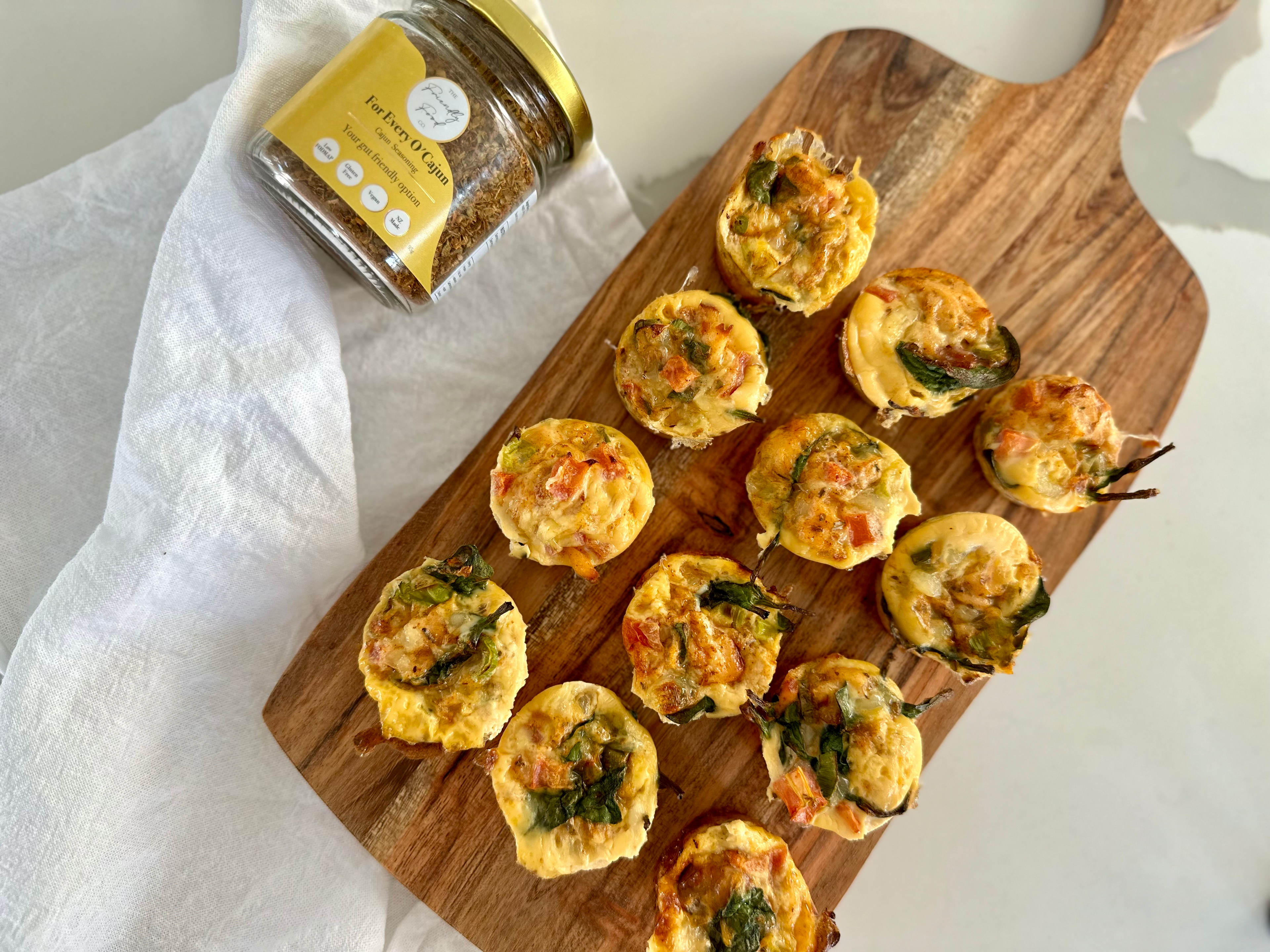 Mini Low FODMAP High Protein Egg and Cottage Cheese Muffin bites on a wooden board with a jar of The Friendly Food Co. Cajun seasoning on a white background