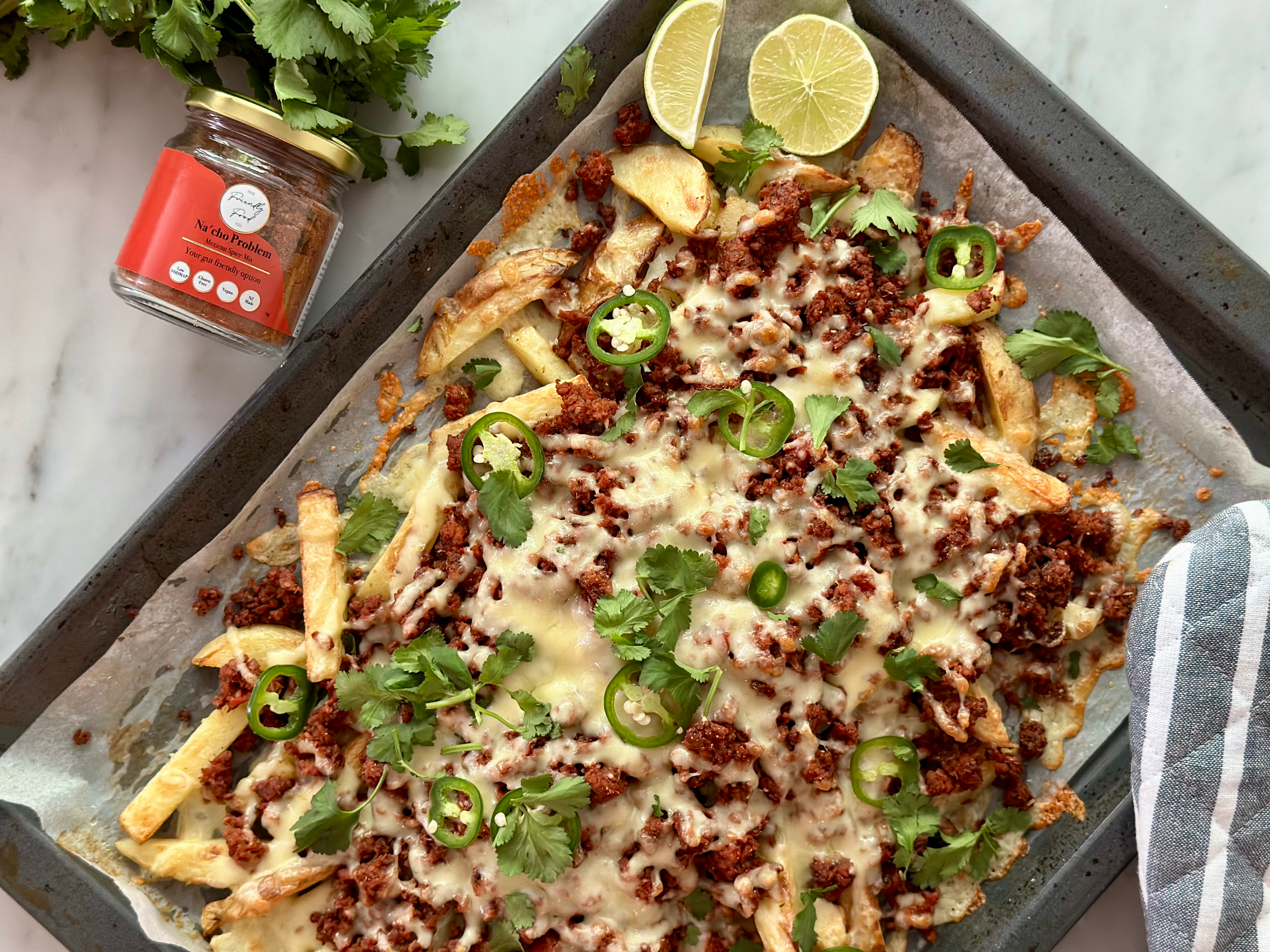 Mexican Loaded Fries with cheese, meat, and jalapeños on a baking tray with a jar of sauce and limes.