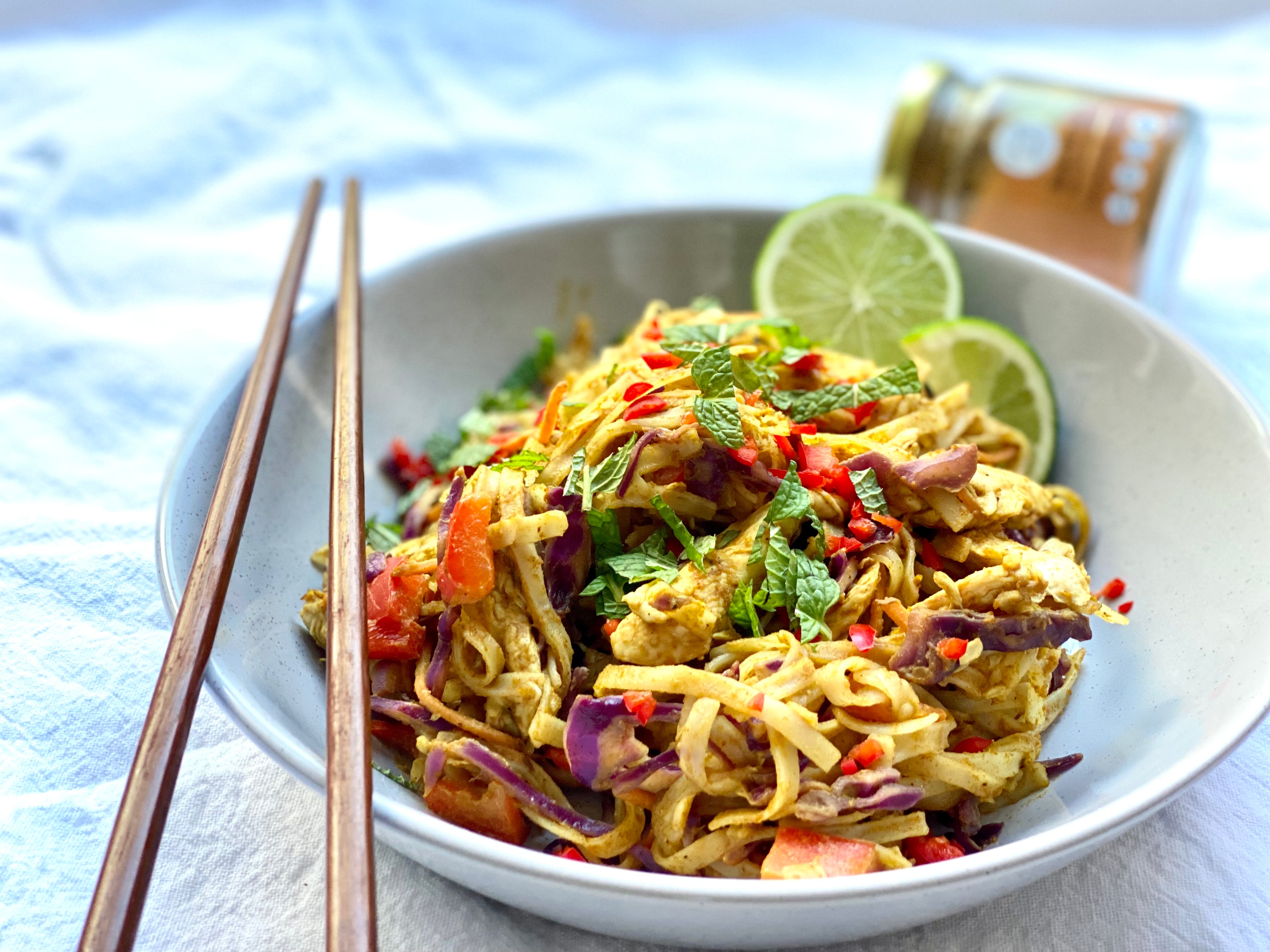 Bowl of Low FODMAP Satay Chicken Noodles with vegetables and lime wedges on a white plate with chopsticks and The Friendly Food Co. Butter Me Up spice mix in the background. 