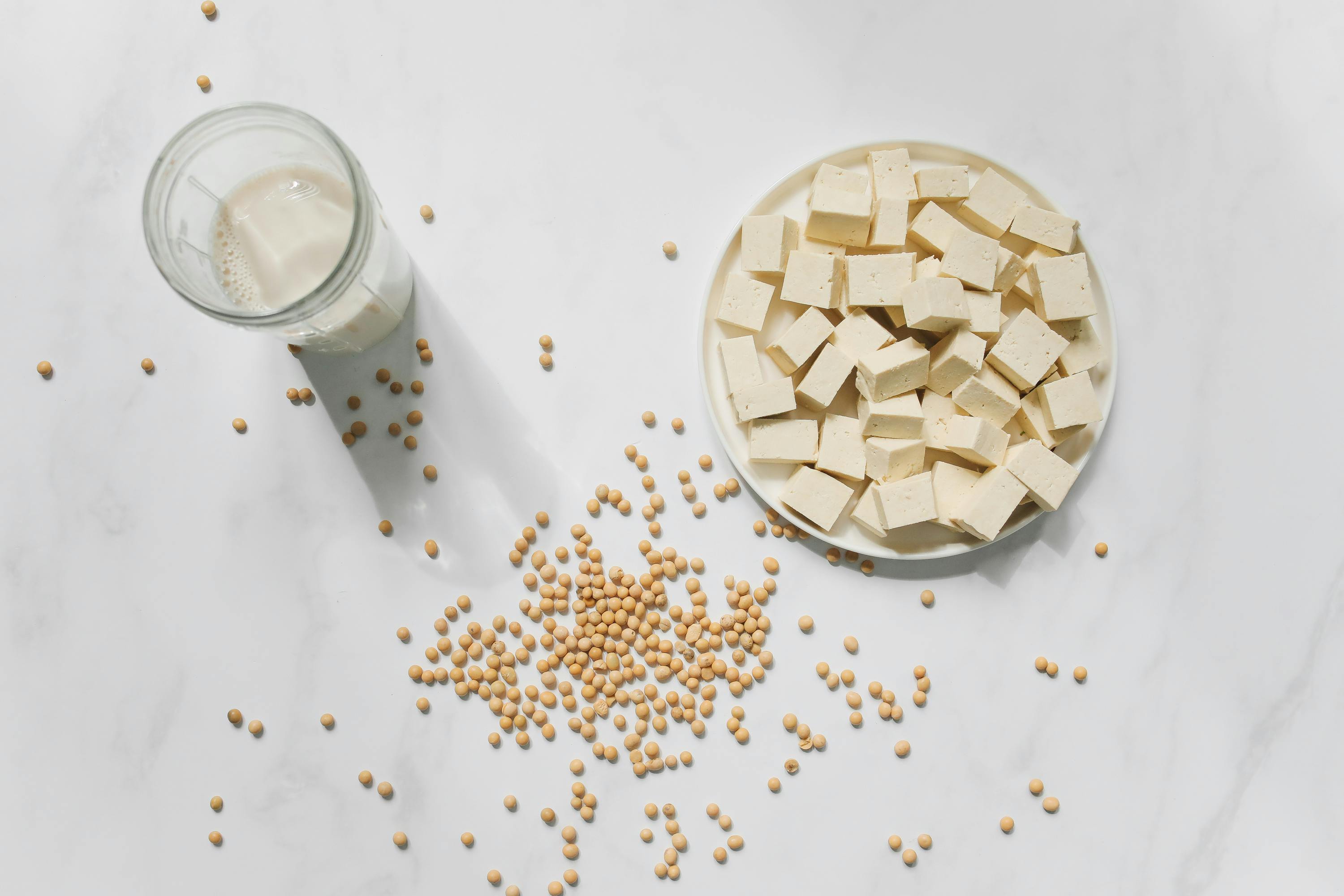Glass of milk, bowl of tofu, and scattered soybeans on a white background
