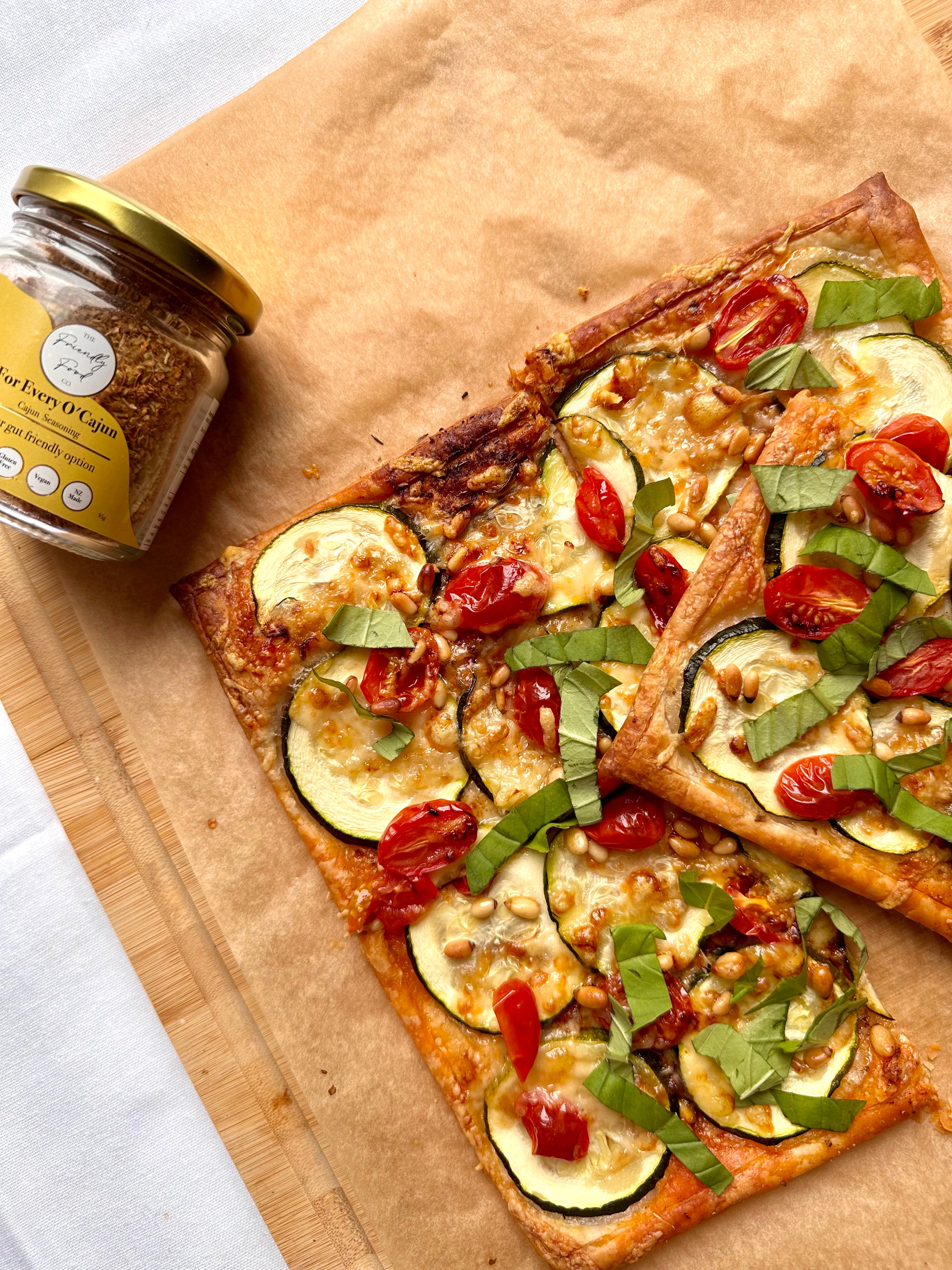 Tomato, Courgette, Gruyere and Basil Tart on a wooden board with a jar of The Friendly Food Co. seasoning.